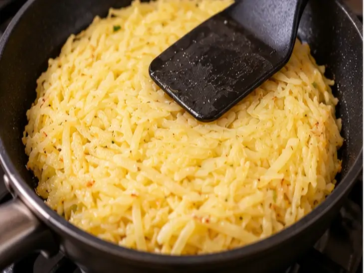Como Moldar Batata Suíça na Frigideira Espátula preta pressionando batata ralada em formato de disco dentro de uma frigideira antiaderente.